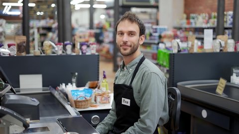 Handsome man in supermarket waiting at cash desk for next customer. Smiling cashier in black apron