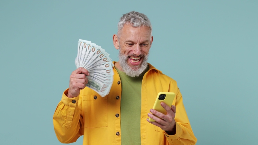 Excited joyful elderly gray-haired bearded man 50s wears yellow shirt using mobile cell phone hold fan of cash money in dollar banknotes isolated on plain pastel light blue background studio portrait