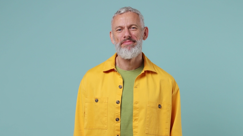 Happy charming elderly gray-haired bearded man 50s wears yellow shirt looking camera wink eye blink isolated on plain pastel light blue background studio portrait. People emotions lifestyle concept