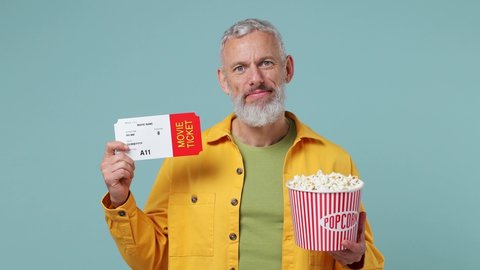 Happy excited satisfied fun elderly gray-haired bearded man 50s wears yellow shirt watch movie film hold bucket of popcorn movie ticket isolated on plain pastel light blue background studio portrait