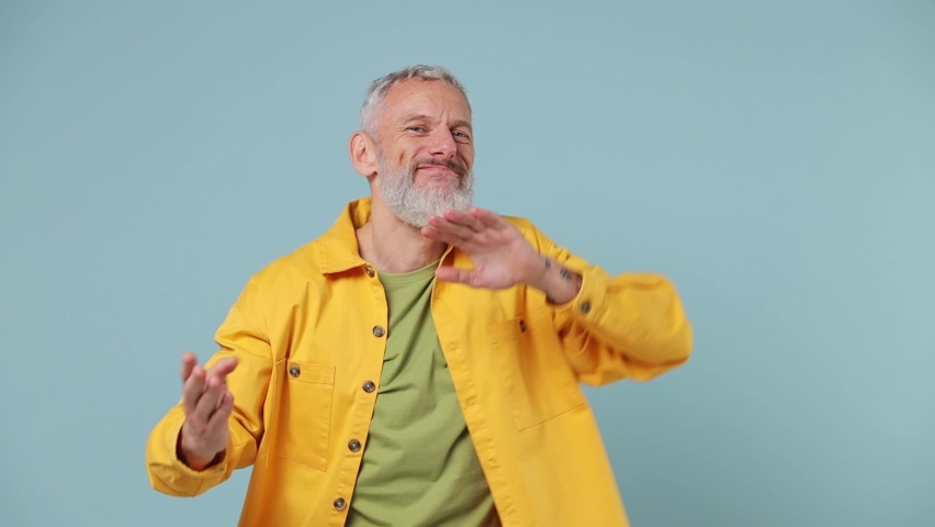 Slow motion fancy vivid elderly gray-haired bearded man 50s in yellow shirt dance fool around have fun gesticulate with hands enjoy relax isolated on plain pastel light blue background studio portrait