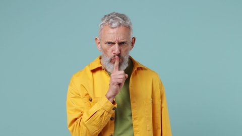 Secret charismatic elderly gray-haired bearded man 50s wears yellow shirt look aside say hush be quiet with finger on lips shhh gesture isolated on plain pastel light blue background studio portrait