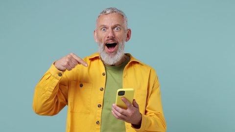 Fun excited surprised elderly gray-haired bearded man 50s wears yellow shirt hold in hand use point finder on mobile cell phone celebrate isolated on plain pastel light blue background studio portrait