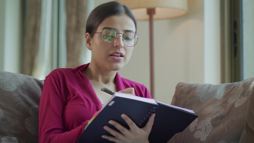 Thoughtful young attractive Indian Asian woman wearing spectacles, writing future business plan or scheduling her next day in her daily journal sitting in an interior home setup. Concept of to do list Royalty-Free Stock Footage #1083857722