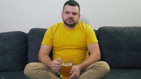 Man in yellow T-shirt is sitting on gray sofa at home in front of the TV and drinking beer in big gulps from glass mug. He is tense and attentive. Relaxing at home, watching TV. Freelance. Alcohol