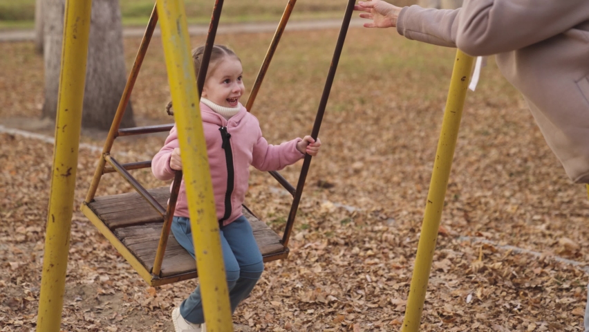 little kid swinging on a swing and laughing while flying up, an autumn playground, a happy family, a mother rolls child in city park, childhood dream to fly, smile at mom on a walk, play with a baby Royalty-Free Stock Footage #1083860185