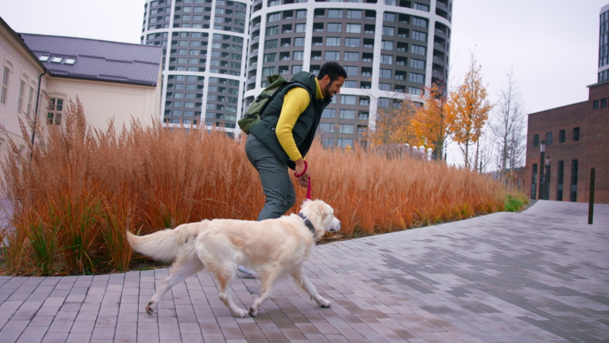 Happy young man running with his dog on leash outdoors in city.