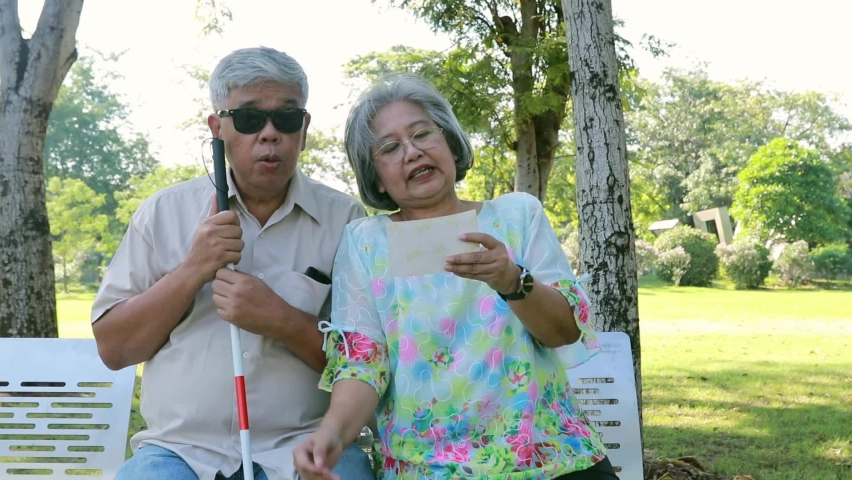 Senior husband and wife couple sit in a chair under the shade of a tree in the park and playfully tell a good story in a photograph as the blind elderly husband sits together and listens intently.