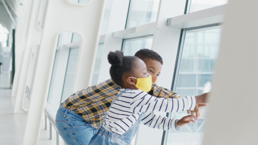 Two African-American children in protective masks spend time waiting for flight. Little brother and sister look at airplanes through large window of airport.