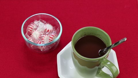 Hand stirring gourmet hot chocolate blend in a green cup near red and white wrapped peppermint candies. Male hand stirring hot chocolate, raising cup out of frame to drink then replacing to saucer. 