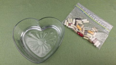 Hand picking up sandwich bag of various nutritional supplements, open and pouring into a heart shape dish and then shaking it around. Nutritional supplements poured into a heart shape dish from a bag