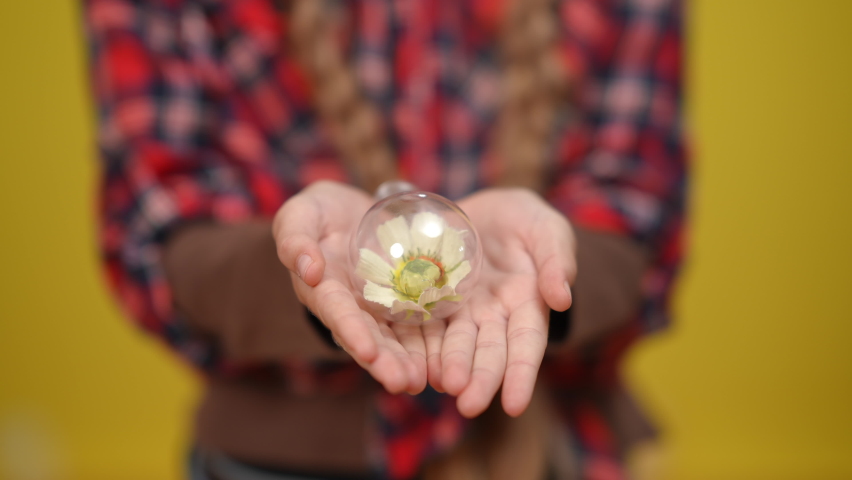 Close-up lightbulb with white flower inside in adolescent Caucasian hands. Unrecognizable teenage girl holding symbol of environmental conservation at yellow background