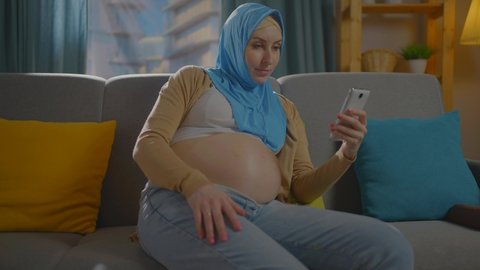 a pregnant muslim woman in scarf is sitting on the couch and experiencing discomfort close up