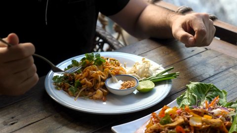 Traditional thai food - pad thai. Man eating fresh food near waterfall at outdoor cafe - thai style fried rice noodle with shrimp, scrambled egg, and bean sprouts. Authentic thai local cuisine dish