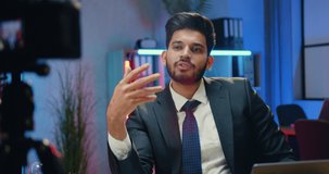 Good-looking confident experienced bearded businessman-blogger in suit recording video for his internet business channel from night lighted office