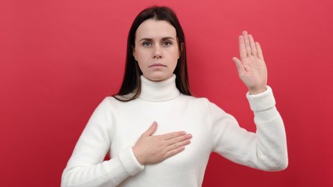 Portrait of serious young female raising hand to take oath, promising to be honest, telling truth, pledging allegiance, wears knitted sweater, posing isolated over red color background wall in studio