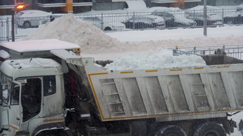 SAINT PETERSBURG, RUSSIA - CIRCA DECEMBER, 2021: Winter snow removal from paths, driveways and parking lots. Tractor with bucket clearing snow and then load them onto dump truck.