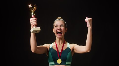 Professional sportswoman winner kissing gold cup, holding golden medal close-up. Woman athlete standing on black background, having first place. Award and victory, winning the championship. 