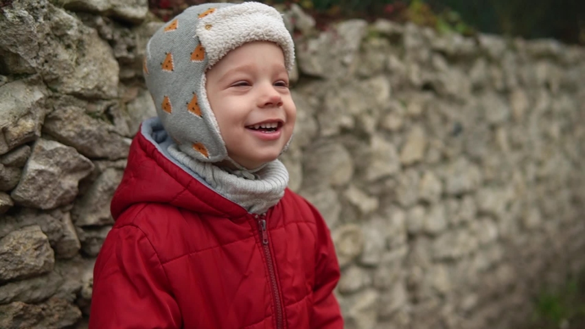 close-up Portrait of European little preschool boy in gray knitted hat red jacket smiling look at camera. Emotionally child laughing smiling posing cover eyes with hands, waves hello outside Autumn