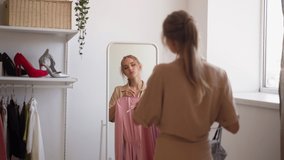 Young blonde woman stands against mirror holding elegant pink and other dresses on hangers to choose for important evening party slow motion