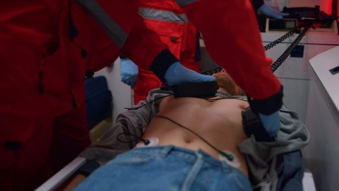 Emergency medical doctors hands using heart defibrillator on patient chest in ambulance car. EMS paramedics providing CPR procedure. Medical assistants rescuing victim with holter monitor device 