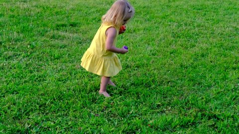 small child, blonde girl 2 years old in yellow dress runs barefoot on green grass in meadow, pick up hidden colored eggs, concept of childhood, traditional activity, fun games for Easter holiday