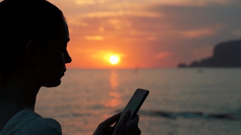 Silhouette of beautiful young woman typing on smartphone in golden lights on beach enjoying sunset sky. summer vacation concept . 4k footage