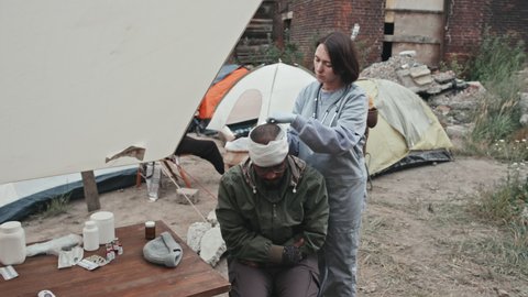 Medium shot of female medical worker bandaging head of wounded man while giving first aid to refugees at tent city