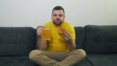Plump young man is sitting on gray sofa and eating big fat burger and drinking beer from large glass mug. He bites the bun in big chunks. Cholesterol, junk food, alcohol, unhealthiness, gluttony fat