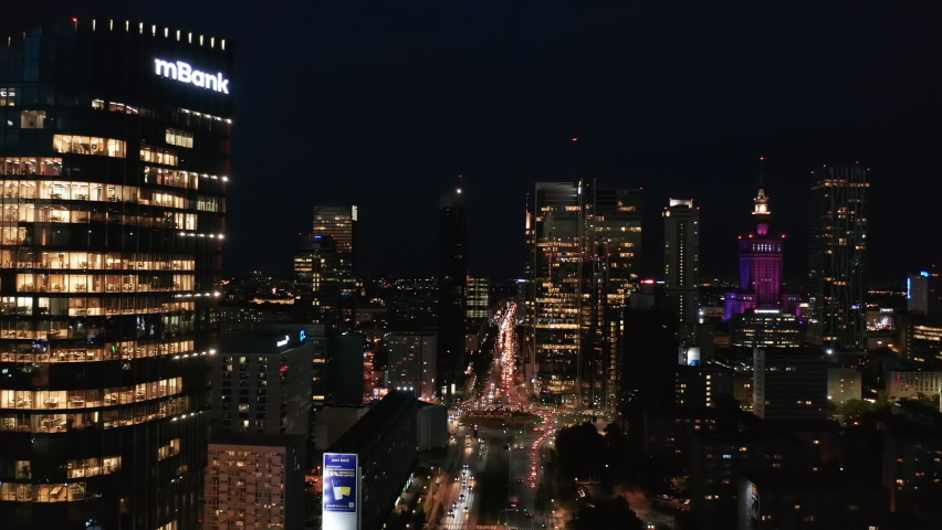 Forwards fly above night city. Heavy traffic on roads and intersection. Illuminated streets and tall commercial buildings around. Warsaw, Poland