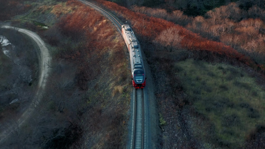 The train rides along the turquoise water in a mountain forest lake with pine trees. Aerial view of the train and lake and green forests. View of the lake between the mountain forest a passenger train