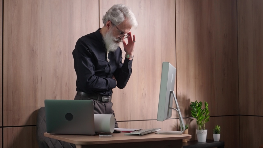 Gray-haired caucasian architect checks drawings of objects and uses a desktop computer for design, a thoughtful engineer checks documents.