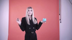Young female putting a coin in piggy bank, saving money concept. Clip. Business blond woman posing at the photo studio with a blue ceramic piggi bank and a coin.