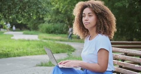 Modern African American woman focused remotely working use laptop sitting on bench at summer park. Female freelancer enjoying distance online communication chatting surfing internet on computer