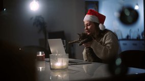 Beautiful young woman in a Christmas hat, taking a video call on her laptop while sitting in the living room with a cute cat in her arms.