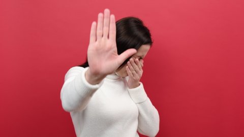 Portrait of young caucasian female grabbing nose with fingers, feeling unpleasant smell, shocked with disgusted fart, wears white knitted sweater, isolated on red color background wall in studio