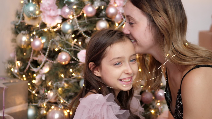 mom and daughter in a pink dress and wrapped in a garland are sitting next to the Christmas tree at home