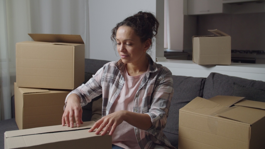 Positive adult woman getting wine glass out of cardboard box, checking it integrity, smiling charmingly, rejoicing successful moving to new home. Many cardboard packages around lady. Housewarming
