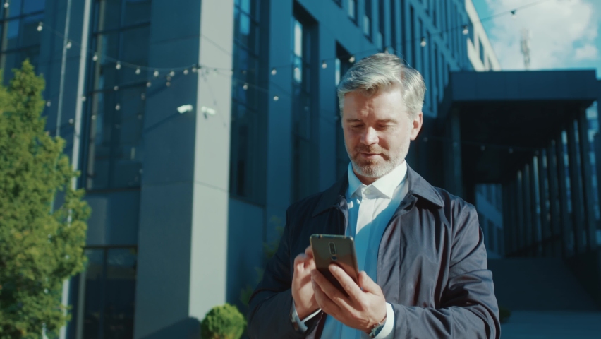 Bearded adult businessman using mobile phone connecting with wife chatting messages walking home after work. Corporate disctrict. Modern city. Urban lifestyle.