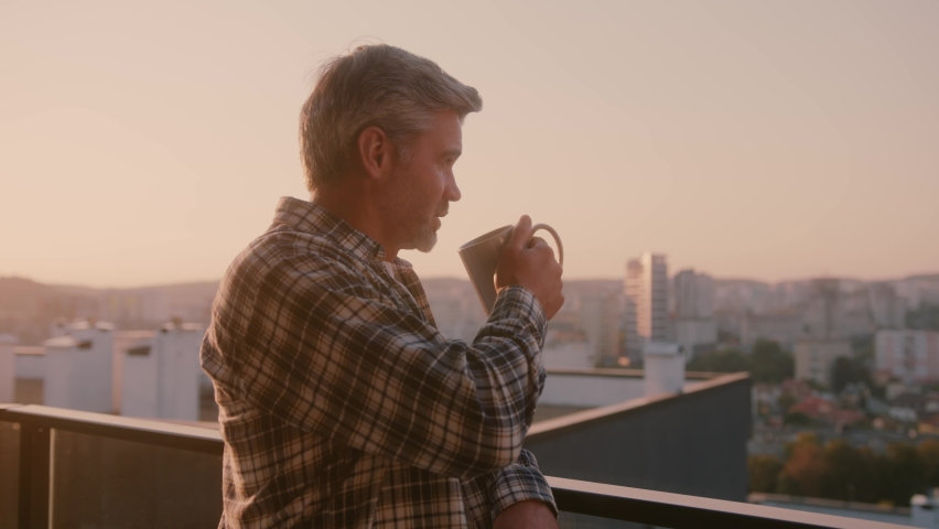 Bearded casual man with cup of coffee enjoying tranquil peaceful sunny morning on balcony apartment. City life. Beautiful scenery.