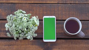 Close-up top view flatlay 4k stock video footage of white cellular cell phone with blank green screen laying on brown wooden table near mug full of tea and simple white bunch of meadow flowers - Powered by Shutterstock - Get 15% off with code: PIKWIZARD15