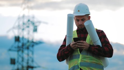 Worker male engineer using mobile phone for checking data while standing against high voltage power towers. Power engineering specialist with smartphone working near electric poles.