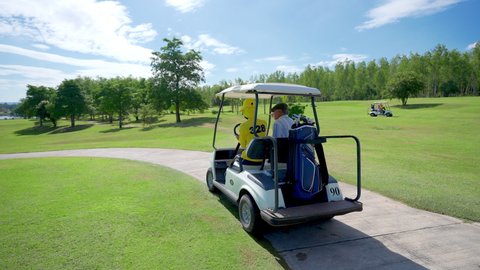 4K Asian senior man with female caddy driving golf cart together on golf course to fairway at country club. Healthy elderly male golfer walking to golfing on the green in summer sunny day