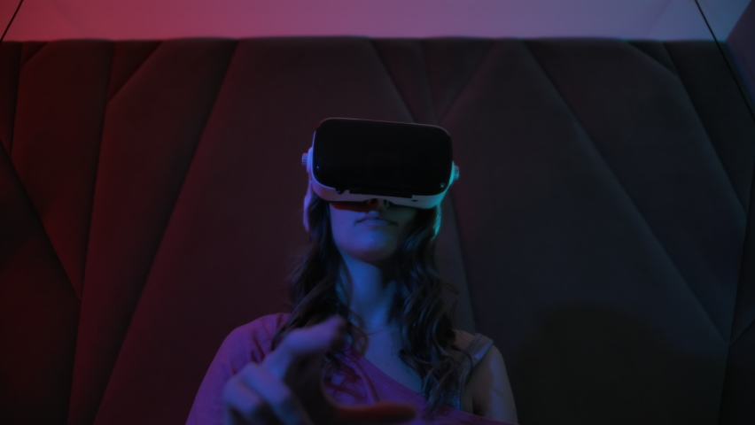 Young Pretty Girl Uses Virtual Reality Glasses. Sitting in Dark Room With Neon Light Playing Games With VR Headset Virtual simulation and training application. New Type Training Royalty-Free Stock Footage #1083922882