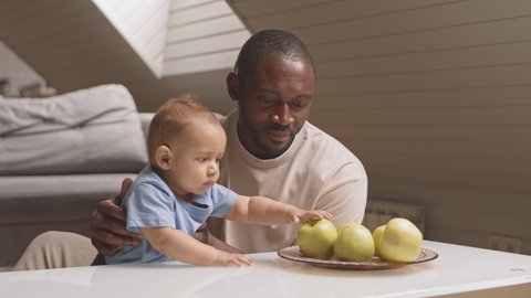 Locked-down of happy African-American father sitting at coffee table at home, holding his cute Biracial baby who taking apple and trying biting it and failing, Caucasian mother joining them
