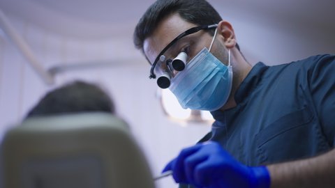 Portrait of professional concentrated Middle Eastern dentist in binocular loupes and face mask examining unrecognizable patient talking in slow motion. Expert orthodontist working in dental clinic