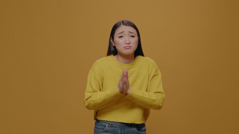 Asian woman doing praying gesture with hands and begging in front of camera, showing desire and expectation sign. Person imploring and asking with palms and body language in studio.