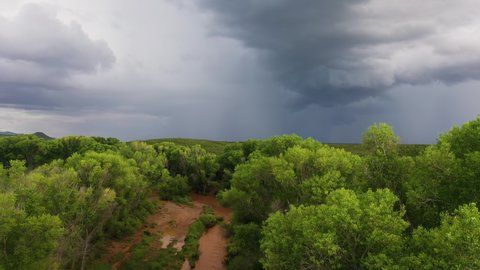 Drone ascend over green cottonwood trees and river with dramatic monsoon clouds