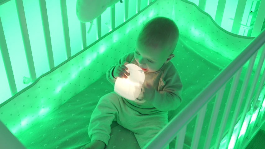 Baby boy sitting in cradle in the evening playing with night light, cute little toddler child in light of baby crib LED night color lighting. High quality 4k footage Royalty-Free Stock Footage #1083941794