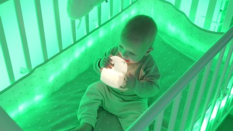 Baby boy sitting in cradle in the evening playing with night light, cute little toddler child in light of baby crib LED night color lighting. High quality 4k footage